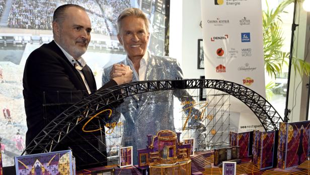 Zwei Männer posieren gemeinsam vor einem detailreichen, bunten Bühnenmodell mit goldenen und violetten Elementen.