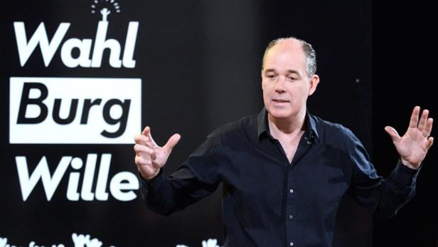 Ein Mann gestikuliert vor einem Wahlplakat mit der Aufschrift „Wahl Burg Wille“.