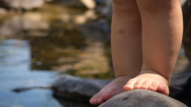 Ein Kleinkind steht barfuß am Rand eines Flusses auf einem Stein, das Wasser ist direkt daneben sichtbar.