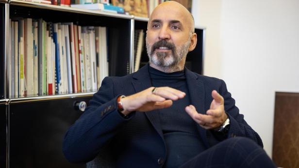 Ein Mann mit Bart und Glatze sitzt vor einem Bücherregal und gestikuliert mit den Händen, als ob er etwas erklärt.