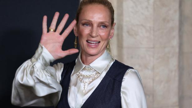Uma Thurman lächelt und winkt mit erhobener Hand in die Kamera.