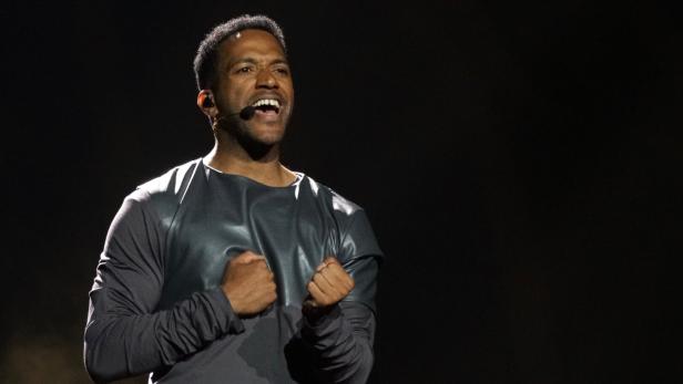 Cesár Sampson mit Mikrofon am Ohr singt mit geballten Fäusten vor dunklem Hintergrund.