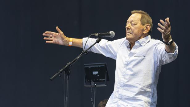 Ein Mann mit ausgebreiteten Armen singt an einem Mikrofon auf einer Bühne vor dunklem Hintergrund.