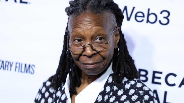 Whoopi Goldberg mit Brille und gemustertem Blazer vor einem Tribeca Film Festival Hintergrund.