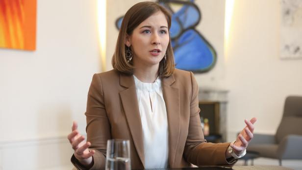 Eine Frau mit braunen Haaren und beigem Blazer spricht gestikulierend an einem Tisch in einem modernen Büro.