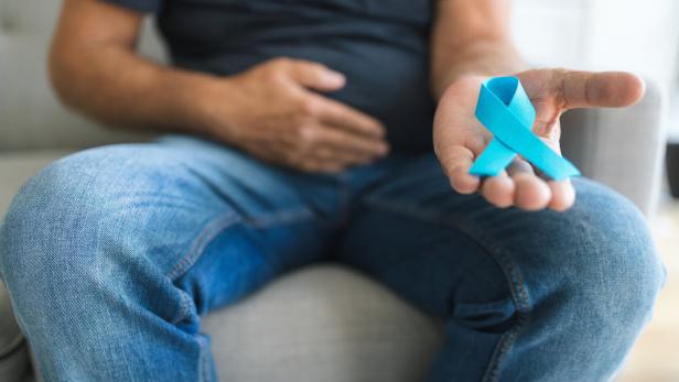 Ein Mann sitzt auf einem Sofa, hält eine hellblaue Schleife in der Hand und fasst sich an den Bauch.