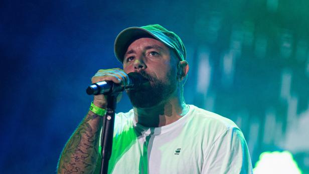 Ein bärtiger Christopher Seiler mit Kappe und Tattoos singt in ein Mikrofon vor blau-grünem Hintergrund.