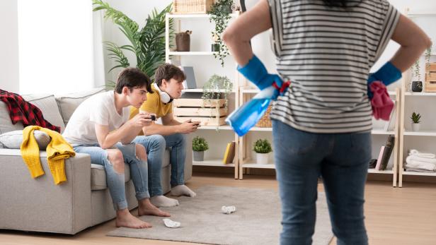 Zwei Jugendliche zocken, während die Mutter die Hausarbeit macht.