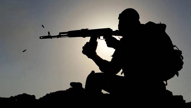 Eine bewaffnete Person in Schutzausrüstung schießt im Gegenlicht, Patronenhülsen fliegen durch die Luft.