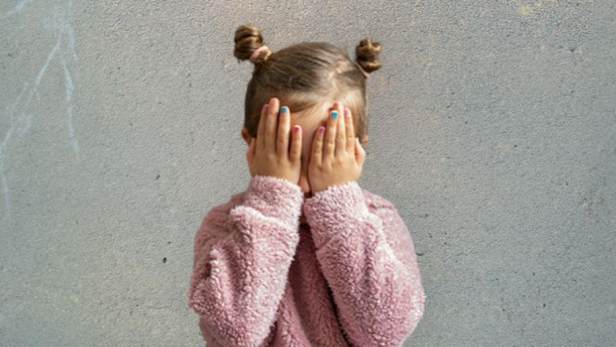 Kind verdeckt das Gesicht mit den Händen vor einer grauen Wand.