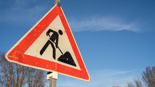 Verkehrsschild mit rotem Rand warnt vor einer Baustelle, im Hintergrund blauer Himmel und Bäume.