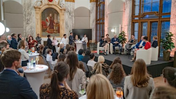 Eine Podiumsdiskussion mit mehreren Teilnehmenden findet in einem prunkvollen Saal vor zahlreichem Publikum statt.
