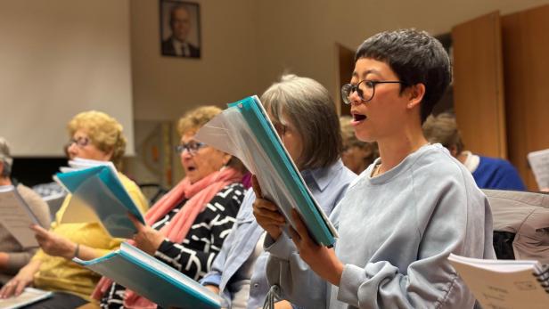 Eine Aufnahme von einer Chorprobe, mehrere ältere Personen sind mit Mappen beim Singen zu sehen. Im Fokus steht eine junge Frau aus Vietnam.