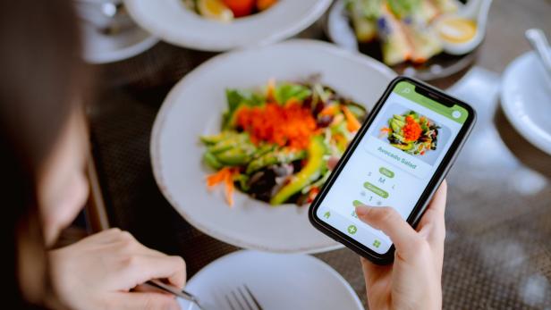 Eine Person sitzt am Tisch mit einem Salatteller und lässt per Handy-App die Kalorien zählen.