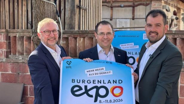Drei Männer im Anzug halten gemeinsam ein großes Plakat für die Burgenland Expo 2026 vor einer Scheune.