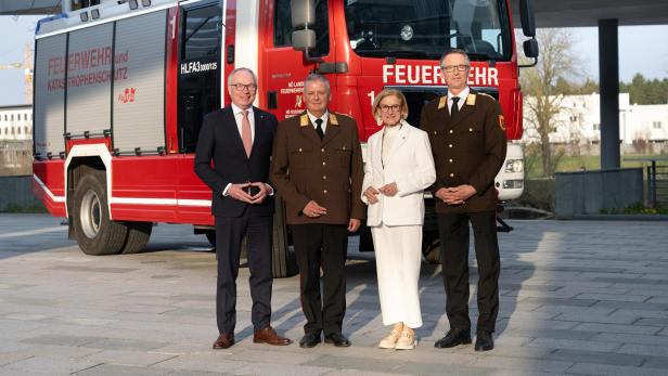 Vier Personen stehen vor einem roten Feuerwehrfahrzeug, drei in Uniform und eine Person im weißen Anzug.