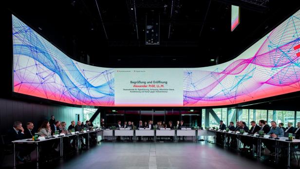 Viele Menschen sitzen an einem großen runden Konferenztisch, über ihnen eine große digitale Anzeigetafel mit buntem Muster.