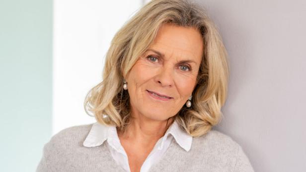 Eine Frau mit blonden Haaren, trägt einen grauen Pullover und Jeans, lehnt an einer Wand.