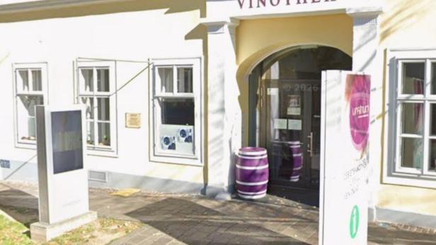 Eingang zu einer Vinothek mit lila Weinfass und Informationsschild vor einem hellen Gebäude.