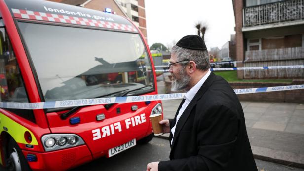 Ein Mann mit Kippa und Pappbecher steht vor einem Feuerwehrfahrzeug, das von Polizeiabsperrband umgeben ist.