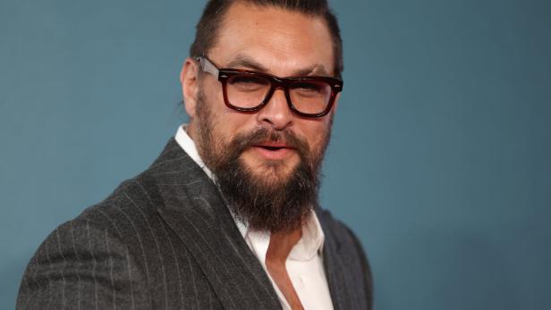 Jason Momoa mit Bart und Brille trägt einen grauen Anzug.