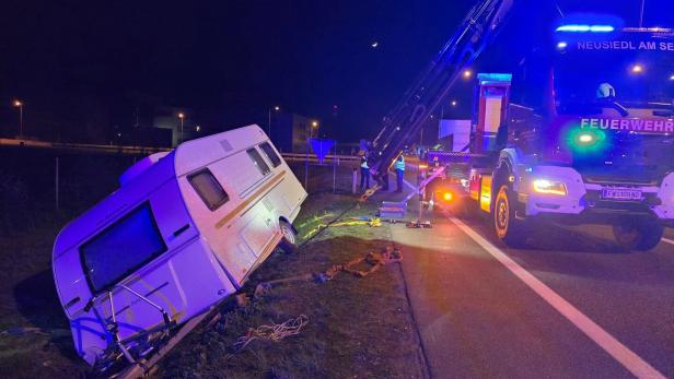 Ein Wohnwagen liegt schräg im Straßengraben, während ein Feuerwehrfahrzeug mit Kran ihn bei Nacht sichert.