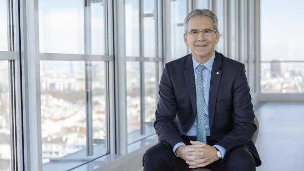Ein weißer Mann sitzt in einem Anzug auf einem Hocker an einer Fensterfront vor der Stadt Wien 