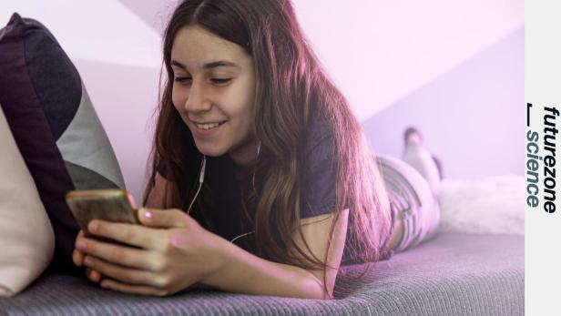 Ein junges Mädchen liegt mit Kopfhörern auf dem Bett und schaut lächelnd auf ihr Smartphone.