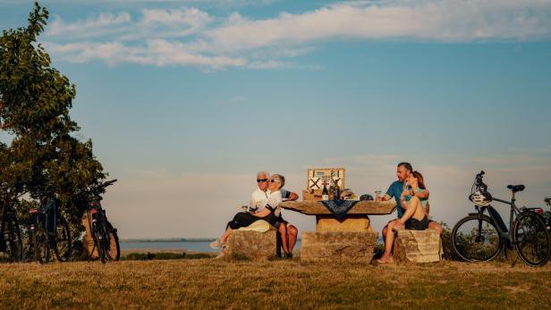 Vier Personen sitzen an einem steinernen Tisch im Freien, trinken Wein und genießen ein Picknick, Fahrräder stehen daneben.