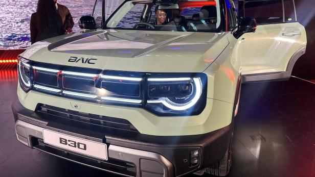 Ein cremefarbener SUV mit geöffneten Türen steht in einer beleuchteten Halle, Menschen unterhalten sich daneben.