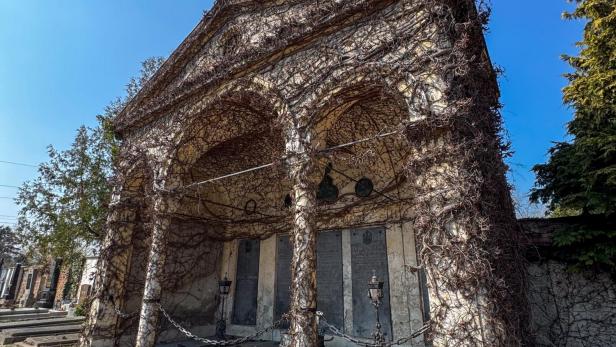 Eine mit Ranken bewachsene, alte Grabstätte mit Säulen und Ketten auf einem Friedhof unter blauem Himmel.