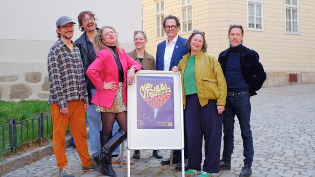 Mehrere bunt gekleidete Menschen, darunter Neubau Bezirksvorsteher Reiter, stehen vor einem Plakatschild auf Pflastersteinen