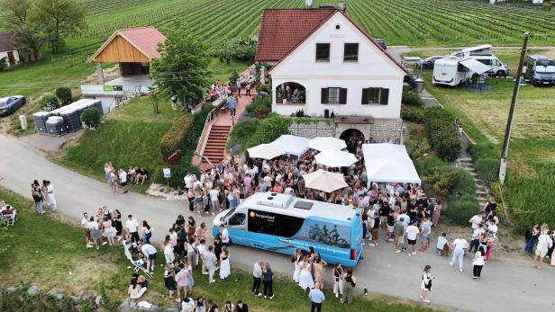 Viele Menschen feiern vor einem weißen Haus mit roten Dachziegeln, umgeben von Weinbergen und mehreren weißen Pavillons.
