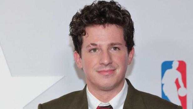 Charlie Puth trägt einen olivgrünen Anzug und eine rot-braun gestreifte Krawatte.