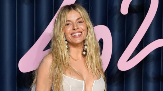 Sienna Miller lächelt in einem weißen, transparenten Kleid vor einem blauen Hintergrund.