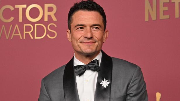 Orlando Bloom trägt einen grauen Anzug mit Fliege und einer Brosche vor einem roten Hintergrund.