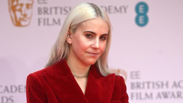 India Rose James mit blonden Haaren trägt einen roten Samtanzug vor dem Hintergrund der BAFTA Film Awards.