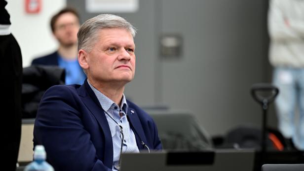 Ein älterer Mann im Anzug sitzt konzentriert an einem Tisch mit Laptop und Wasserflasche.