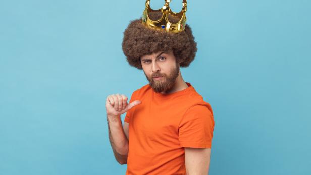 Mann mit Lockenperücke und goldener Krone trägt ein orangefarbenes T-Shirt und zeigt mit dem Daumen auf sich selbst.