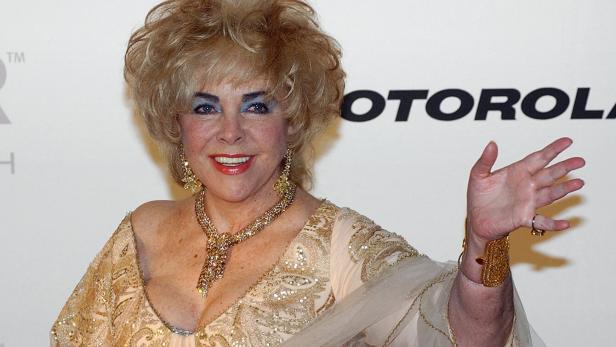 Elizabeth Taylor in einem goldenen Kleid mit Schmuck winkt vor einem Motorola-Logo.