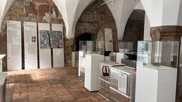 Ausstellung in einem Raum mit Gewölbedecke, Informationstafeln und Vitrinen auf weißen Sockeln.