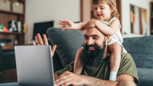 Ein Mann lernt mit einem Laptop zu Hause, während ein kleines Kind auf seinen Schultern sitzt und beide lächeln.