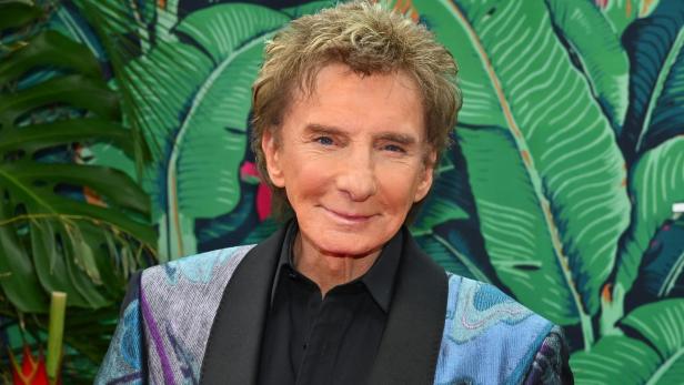 Barry Manilow lächelt vor einem Hintergrund mit tropischen Blättern.