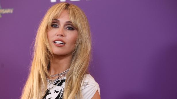 Miley Cyrus mit blonden Haaren und Halsketten vor lila Hintergrund.