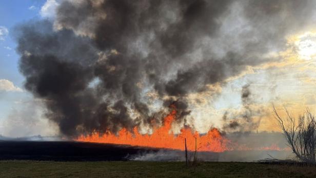 Großes Feuer mit dichten schwarzen Rauchwolken breitet sich auf einem Feld aus, während die Sonne am Himmel steht.
