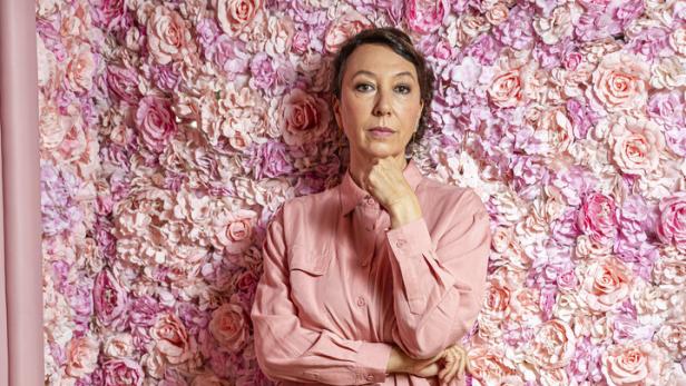 Frau in rosa Outfit steht vor einer Wand aus rosa und pinken Kunstblumen und schaut nachdenklich.