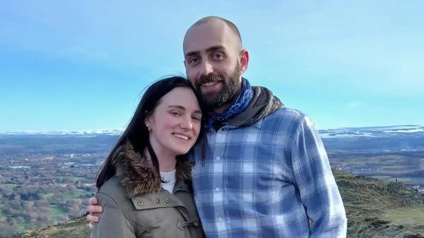 Ein junges Paar steht lächelnd auf einem Hügel mit weiter Landschaft im Hintergrund.