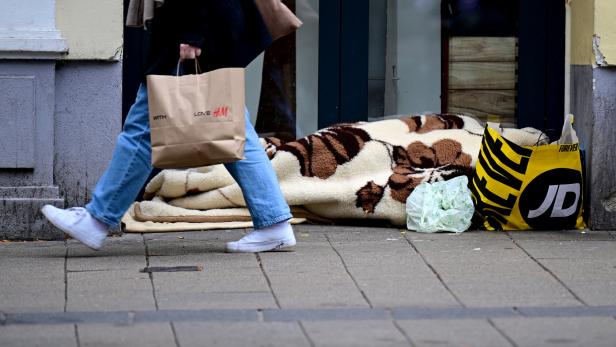 Eine Person liegt unter einer Decke auf dem Gehweg, daneben Einkaufstüten, während jemand vorbeigeht.