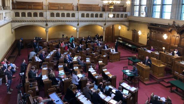 Der Gemeinderatssaal mit vielen Personen in Bänken, einem Rednerpult und einem großen Kronleuchter.