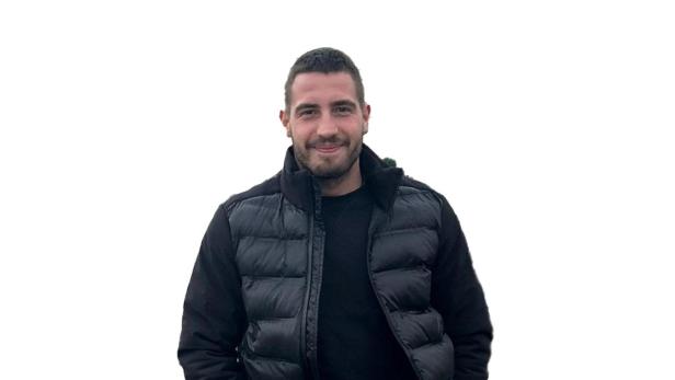 Der von der Wiener Polizei gesuchte 26-jährige Verdächtige steht in voller Körperansicht mit dunkler Jacke und Jeans vor hellem Hintergrund.